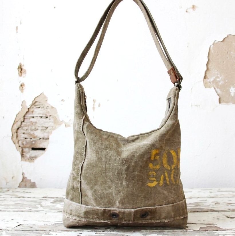 Vue de face d'un sac bandoulière éco-responsable pour femme, suspendu dans un atelier artisanal français. Le sac est en toile militaire recyclée robuste, de couleur vert olive, avec des écritures d'origine en jaune. Il a une sangle réglable et est posé au-dessus d'une table en bois patiné, devant un mur en plâtre écaillé.