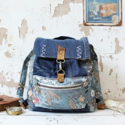 Sac à dos artisanal pour femme en denim bleu recyclé et chutes de tissu tapisserie à motifs fleurs et écailles, style vintage bohème, sur fond blanc rustique.
