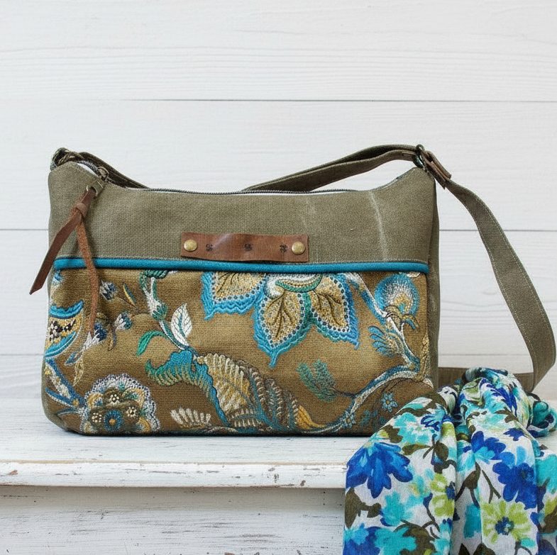 Une femme, portant un haut en coton blanc brodé et un jean bleu, tient un smartphone au-dessus d'un sac bandoulière en toile d'inspiration vintage et bohème de couleur olive, dont elle ouvre la fermeture éclair. Le sac présente un empiècement inférieur avec un motif floral brodé complexe dans des teintes d'azur, d'or et de crème. Il a une bordure en tissu azur et une petite languette de tirage en cuir. L'arrière-plan est un mur blanchi à la chaux usé avec une fenêtre, créant une atmosphère rustique et artisanale.